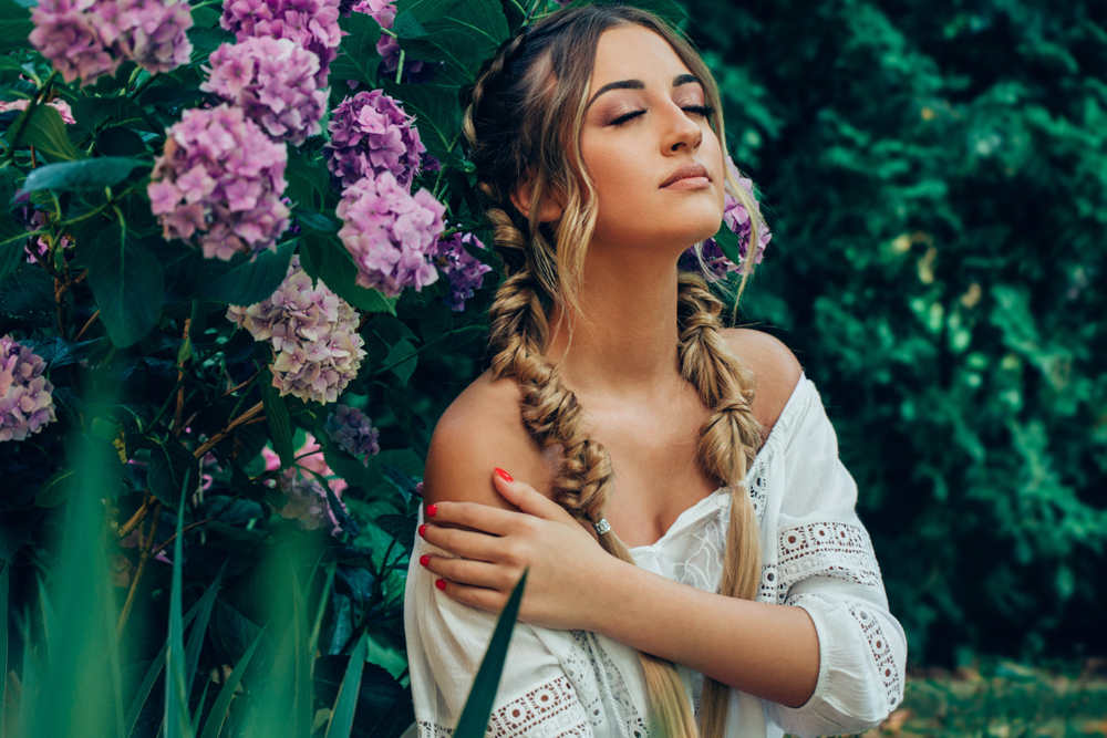 Meisje sluit haar ogen en raakt haar schouder aan buiten met bloemen achter haar die een bubble braid kapsel dragen met fishtail vlechten