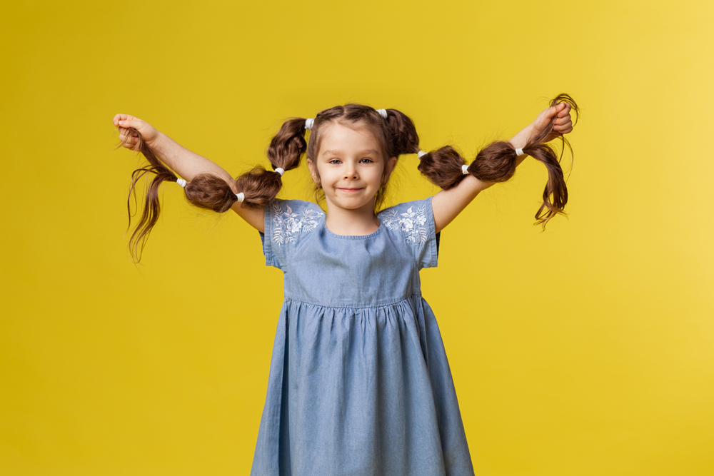 Het meisje staat in blauwe kleding voor gele achtergrond die lange bellenvlechten omhoog houdt