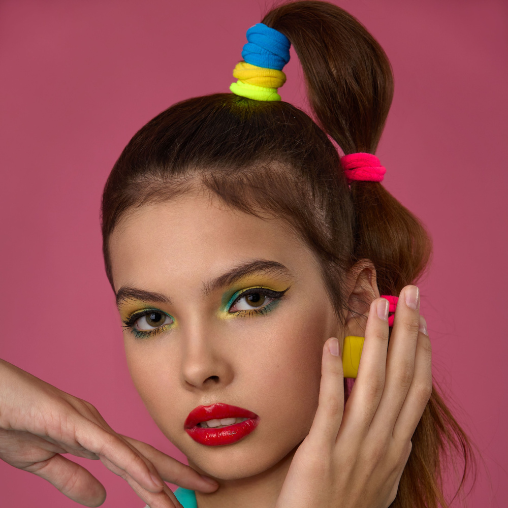 Close up view van brunette vrouw met regenboog gekleurde elastieken die jaren '80 bubble braid kapsel draagt voor roze muur met hand op haar gezicht