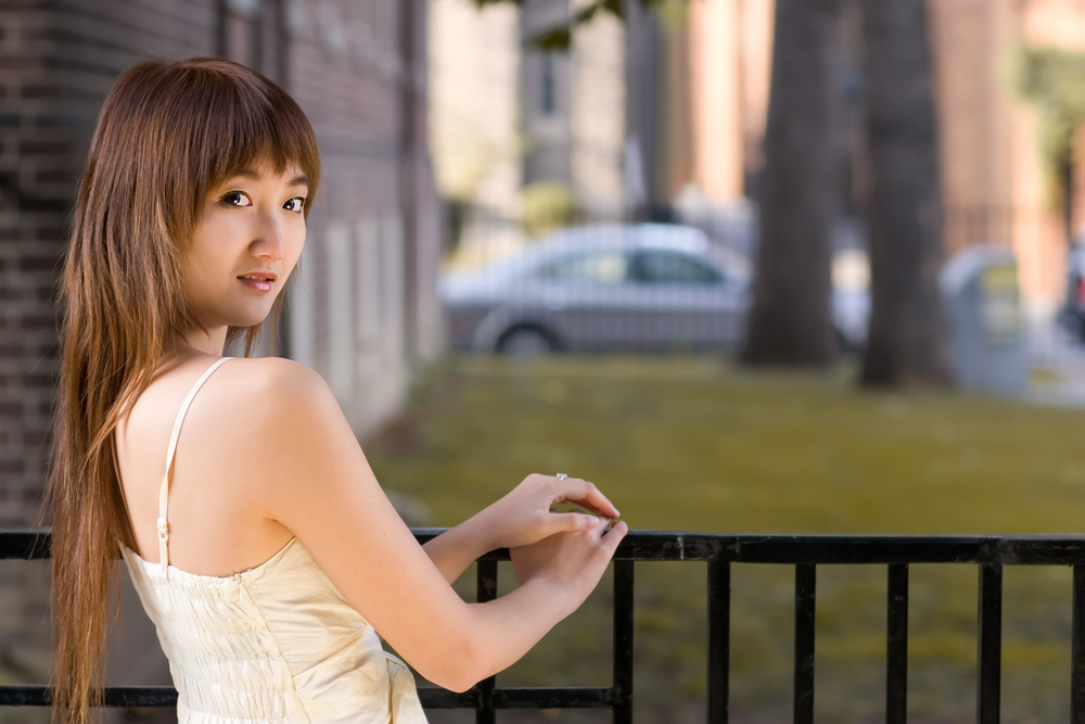 Jonge Aziatische vrouw staat bij reling buiten en kijkt over haar schouder met witte jurk die haar versie van lange shagkapsels toont.