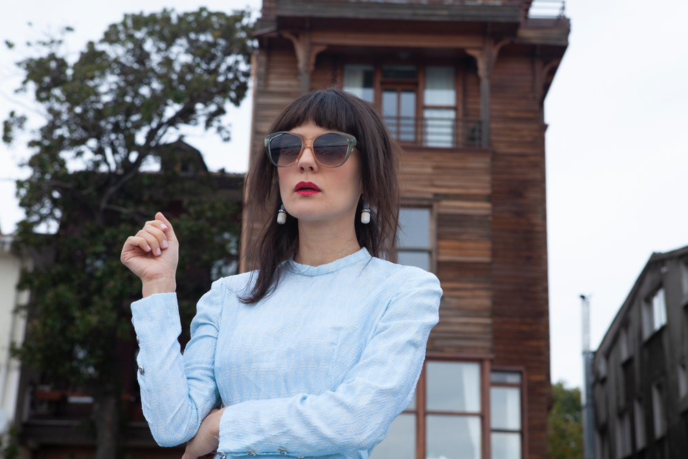 Brunette vrouw draagt grote zonnebril voor houten gebouw met blauwe blouse om voorbeelden van lange shag kapsels te tonen