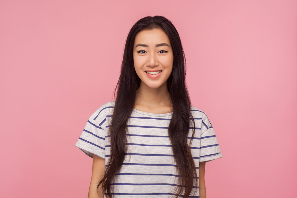 Jong Aziatisch lang haar model met gestreept hemd dat voor roze muur glimlacht