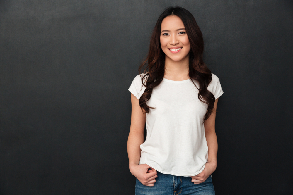 Gelukkige Aziatische vrouw met middenruglengte haar draagt een wit overhemd en jeans voor een donkergrijze achtergrond met handen door haar riemlussen