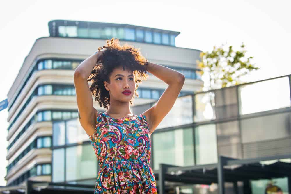 Afro-Amerikaanse vrouw met krullend haar dat is gehydrateerd met cederhoutolie en de stad met haar armen omhoog staat te spelen met haar haar.