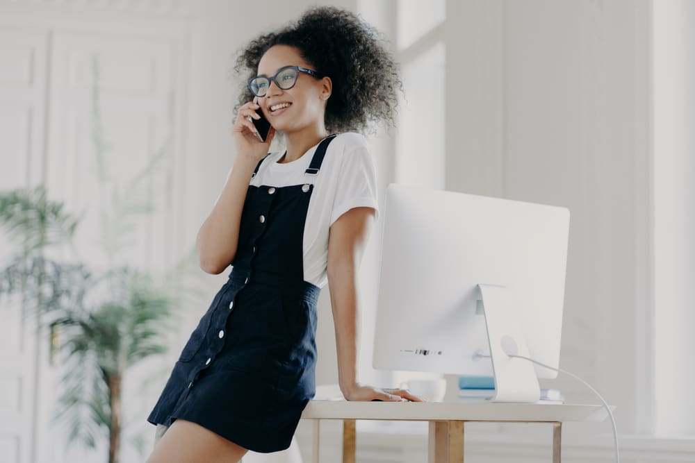 Vrouw met krullend haar pratend aan de telefoon op kantoor die Earl Gray-thee drinkt met bergamot in een zwarte jurk en wit t-shirt. 