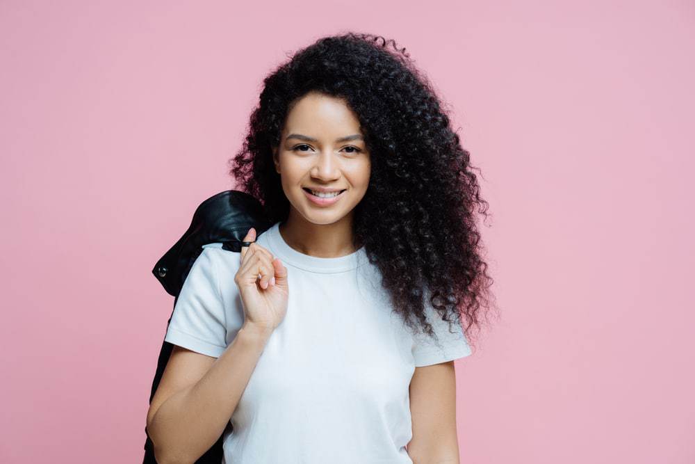 Etnisch Afro-Amerikaanse vrouw met krullend haar die een wit t-shirt draagt en een zwart leren jasje over haar schouder draagt. 