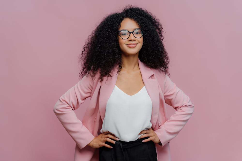 Zakenvrouw met gemengd ras in een roze blazer, wit t-shirt en zwarte bril met zwart krullend haar. 