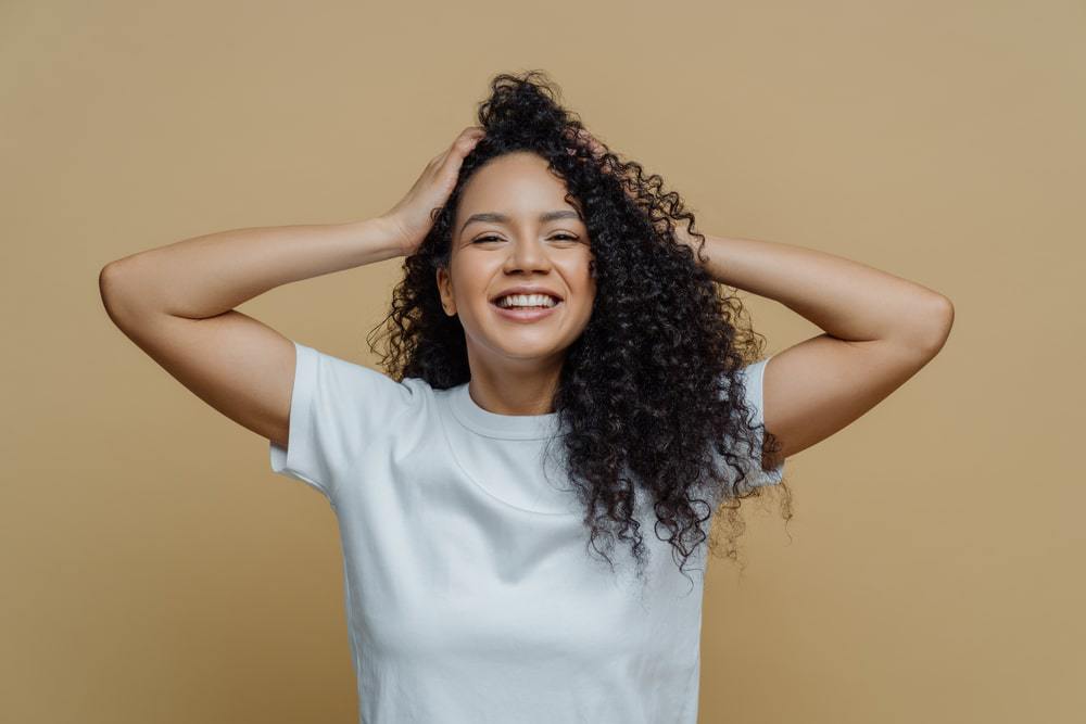 Gelukkig zwart meisje met curly haar vasthouden haar lokken die waren gehydrateerd met bergamotolie terwijl het dragen van een wit t-shirt.