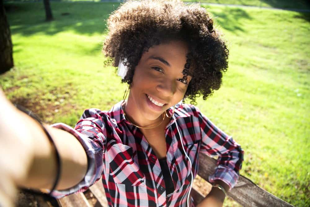 Close-up portret van glimlachende jonge aantrekkelijke Afro-Amerikaanse vrouw die een smartphone vasthoudt, selfiefoto maakt en in het park staat met wazige planten en gras op de achtergrond.