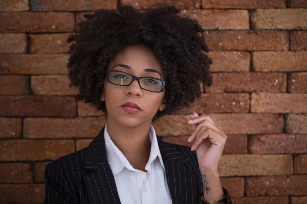 Mooie Afro-Amerikaanse zakenvrouw met bril op kantoor die voor een bakstenen muur staat.