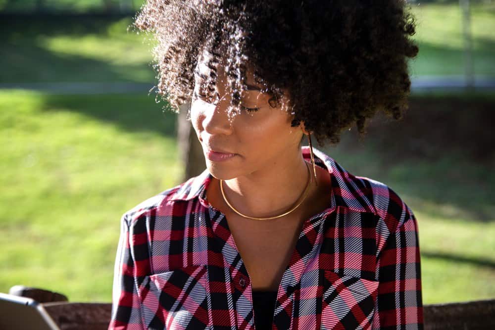 Jonge mooie Braziliaanse vrouw met zwart krullend Afro-haar met tablet, terwijl ze buiten op de houten bank in een park zit