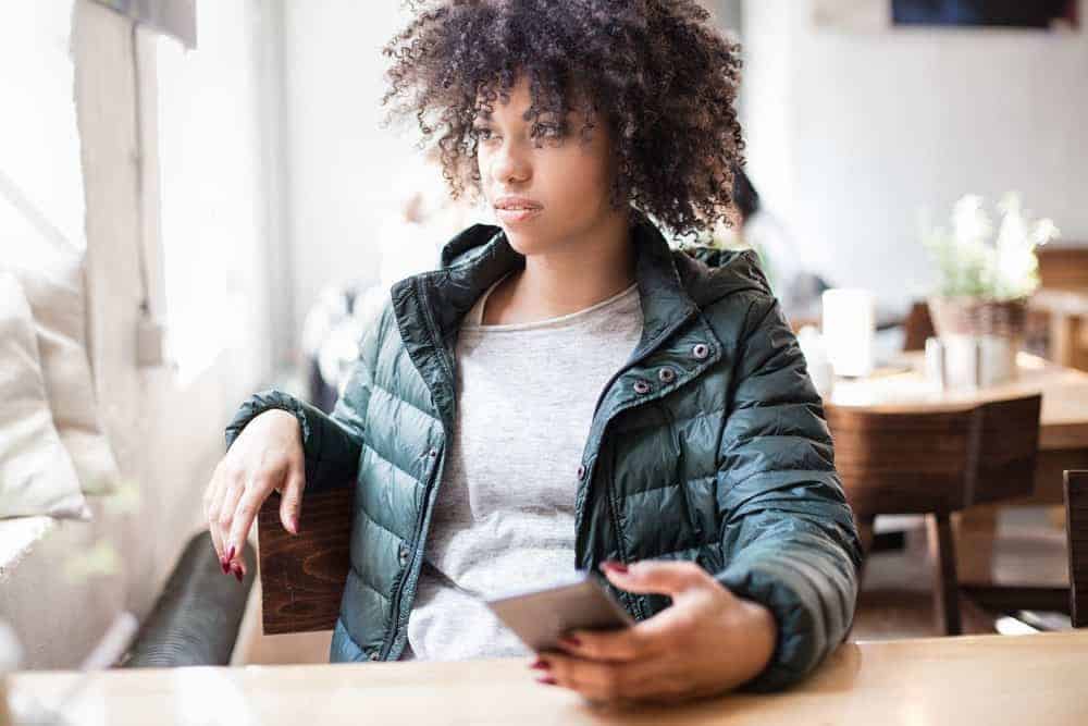 Mooie Afro-Amerikaanse vrouw die aan een tafel zit met een groen jasje, met behulp van een mobiele telefoon met krullend haar.