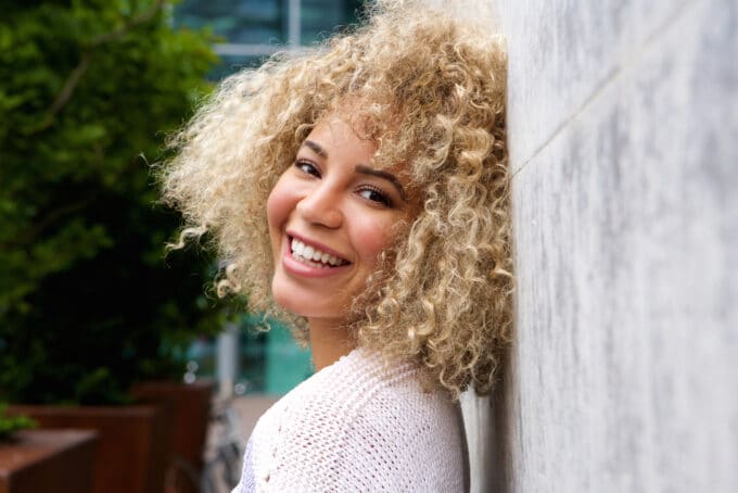 Zwarte vrouwen met type 3B krullend haar geverfd blond met semi-permanente haarverf leunend op een stenen muur.