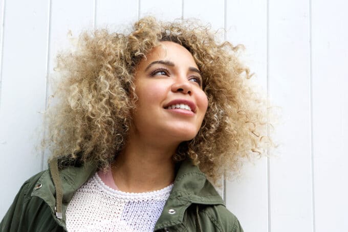 Mooie zwarte vrouw die in direct zonlicht staat met blond type 3b krullend haar in een groene jas en witte trui.