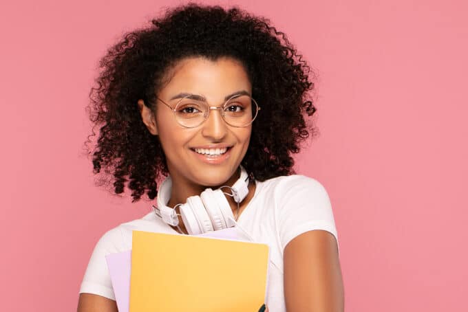 Leuke zwarte vrouw met ronde bril, roze lippenstift, krullende, witte Dr. Dre-koptelefoon en een geel notitieboekje.