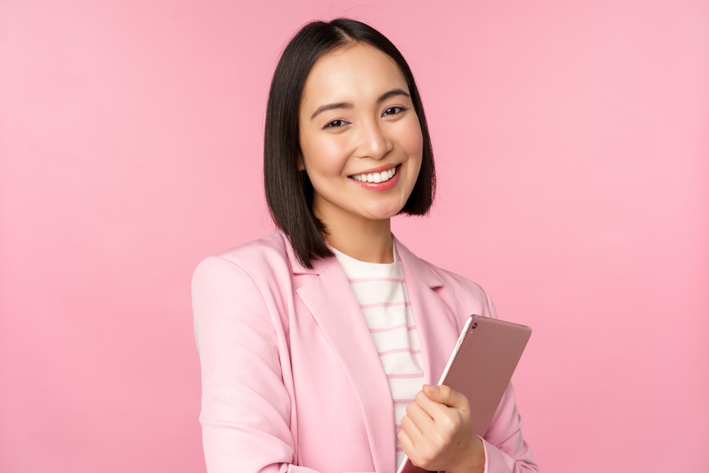 Glimlachende vrouw met donker haar houdt een tablet in roze blazer voor roze muur om haar korte kapsel op een hartvormig gezicht te tonen