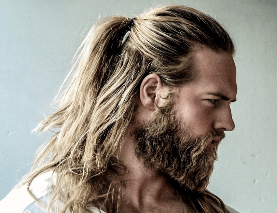 Viking kapsels: Man met lang donkerblond haar gestyled in half opgetrokken paardenstaart met stoere baard, gekleed in wit overhemd