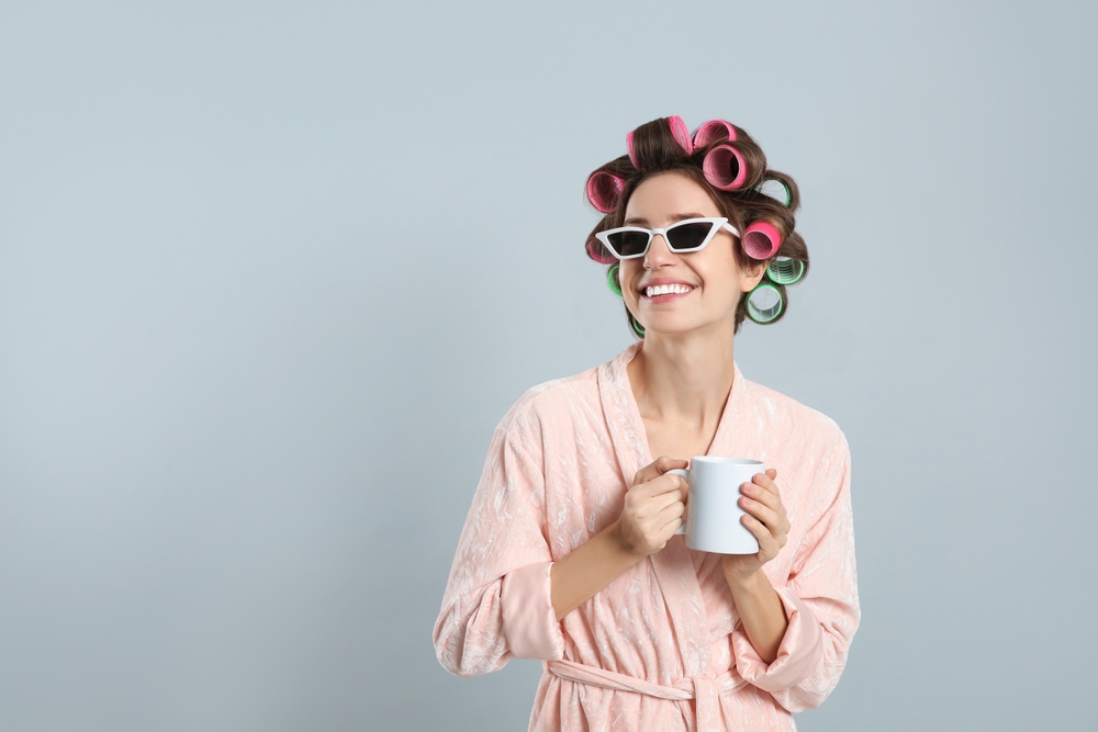 Vrouw in badjas draagt krulspelden in haar haar voor gekke haardag ideeën met witte zonnebril en een mok terwijl ze glimlacht voor grijze muur