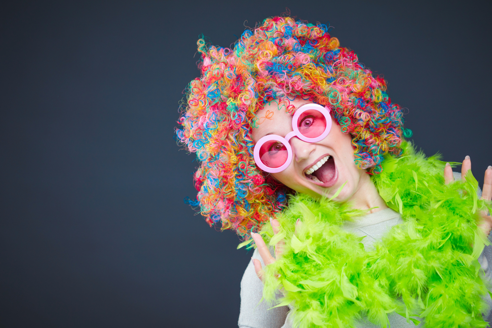 De vrouw draagt een kleurrijke afro krullende pruik met groene veerboa voor donkere achtergrond terwijl het glimlachen in grote roze zonnebril