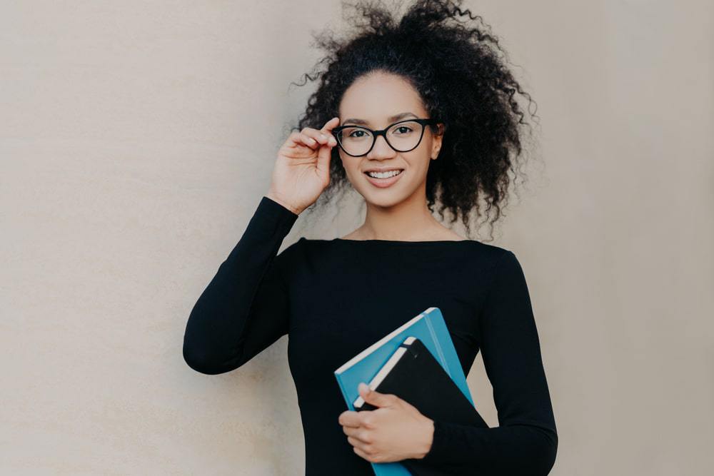 Leuke zwarte vrouw met kroeshaar met een zwarte trui, leesbril en twee haarverzorgingsdagboeken. 