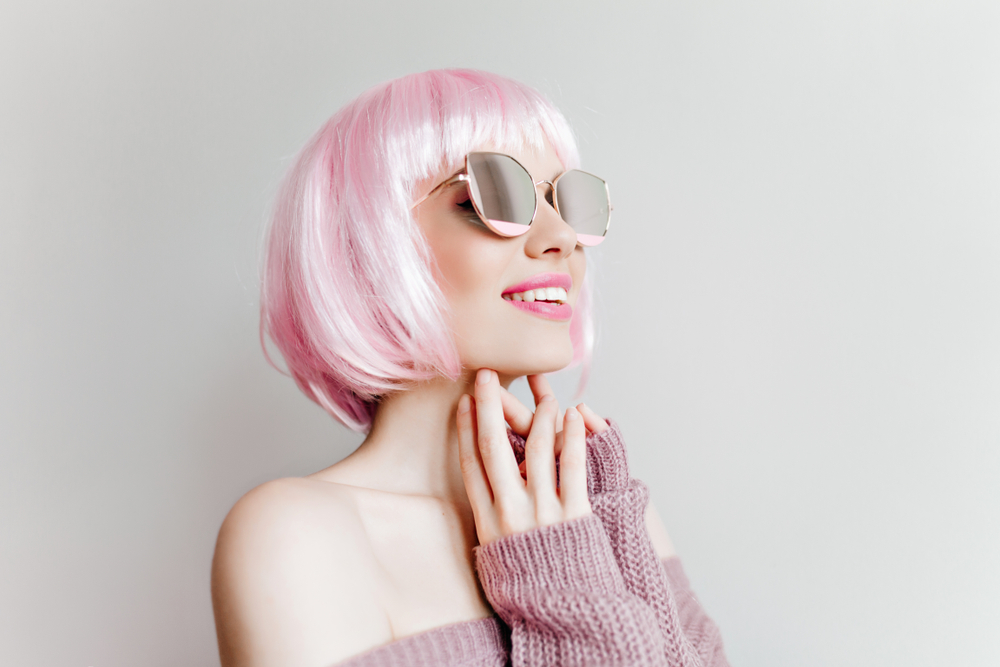 White Taffy Pink Blonde afgebeeld met haar handen onder haar kin lachend en opkijkend door haar zonnebril.