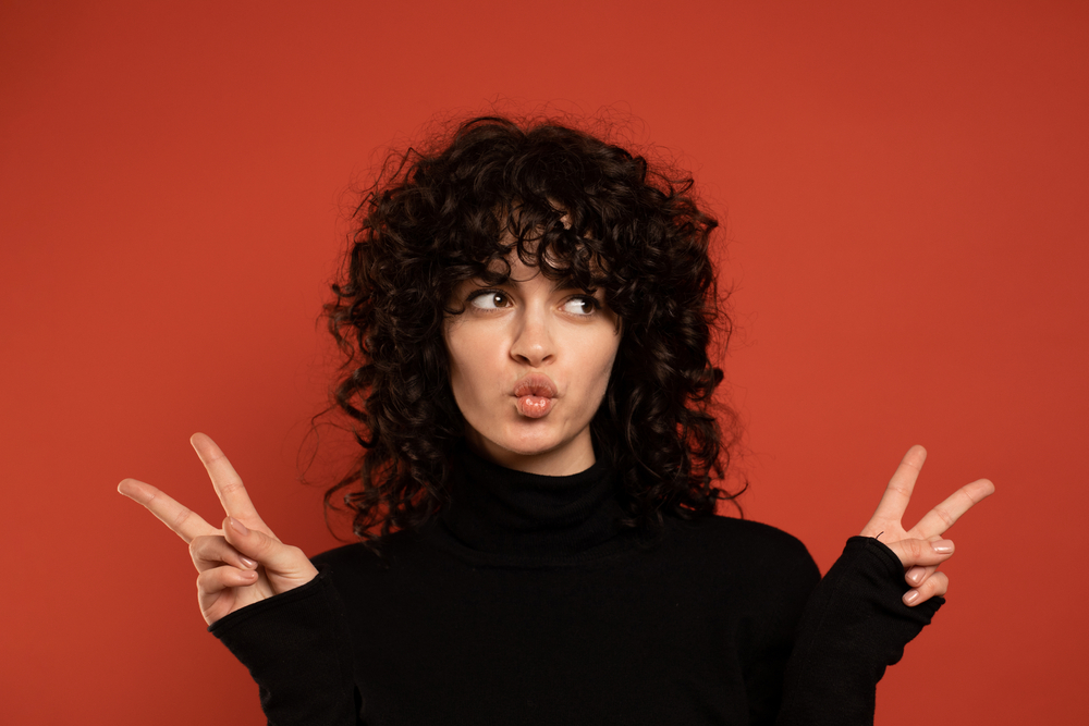 Curly Shag With Wispy Bangs op een vrouw met een 80's vibe die het vredesteken geeft in een rode kamer.