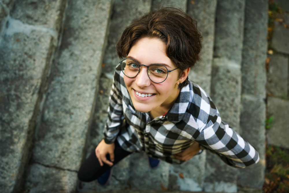 Mooie tomboy in een geruit overhemd en een ronde bril glimlacht en kijkt op naar de camera voor een stukje over de beste tomboy korte pluizige kapsels