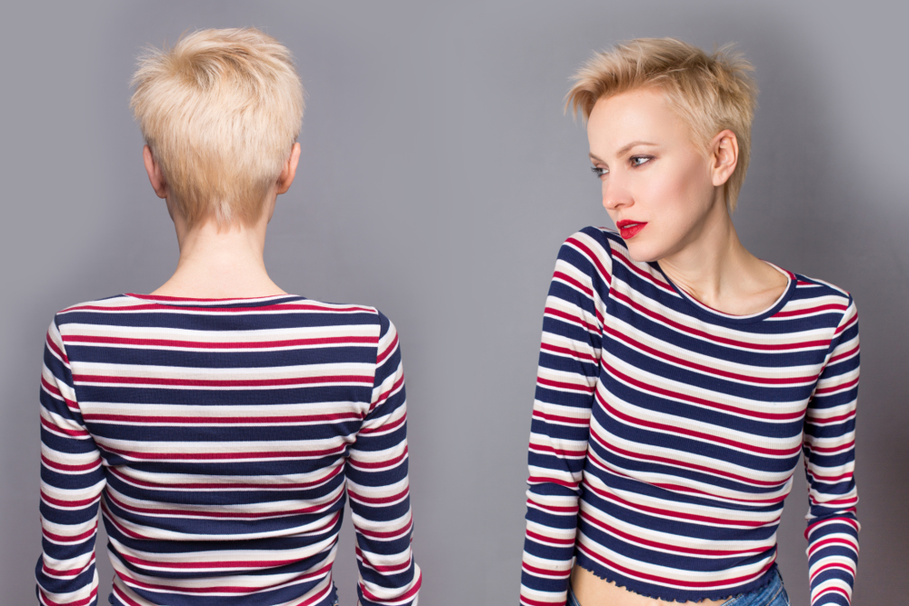 Blonde vrouw met kort haar draagt een gestreept rood en blauw shirt voor een grijze muur om te laten zien hoe ze een korte pixie cut stylt.