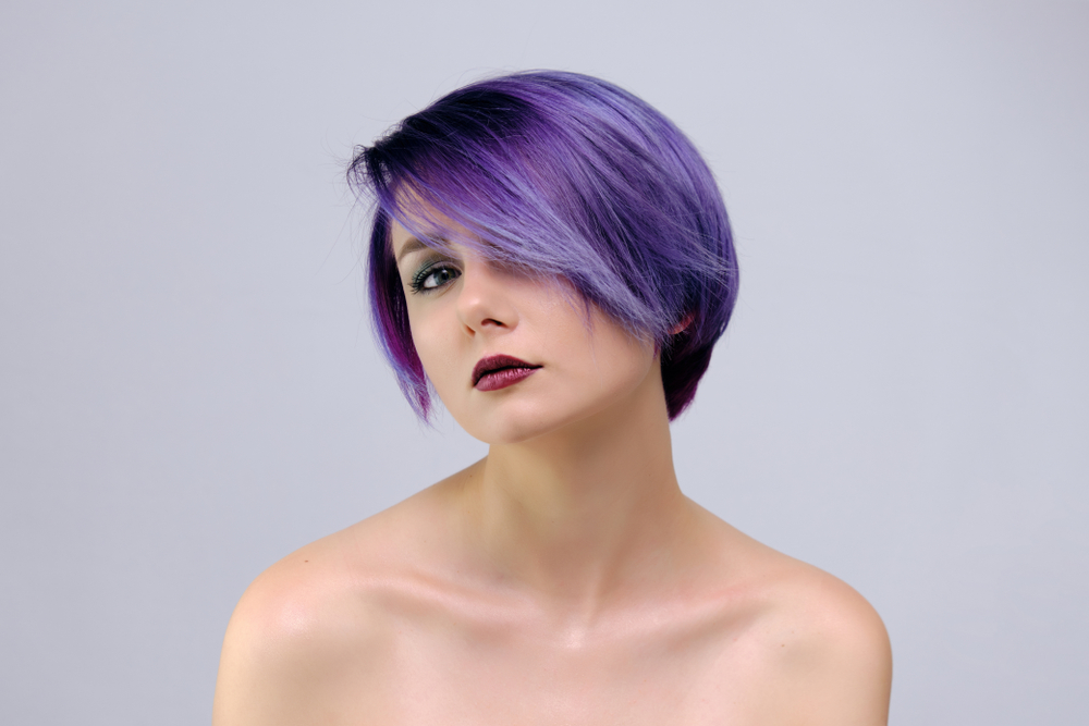 Vrouw met blote schouders modelleert haar lavendel kleur met een bixie cut en lichtere paarse highlights voor een grijze muur.