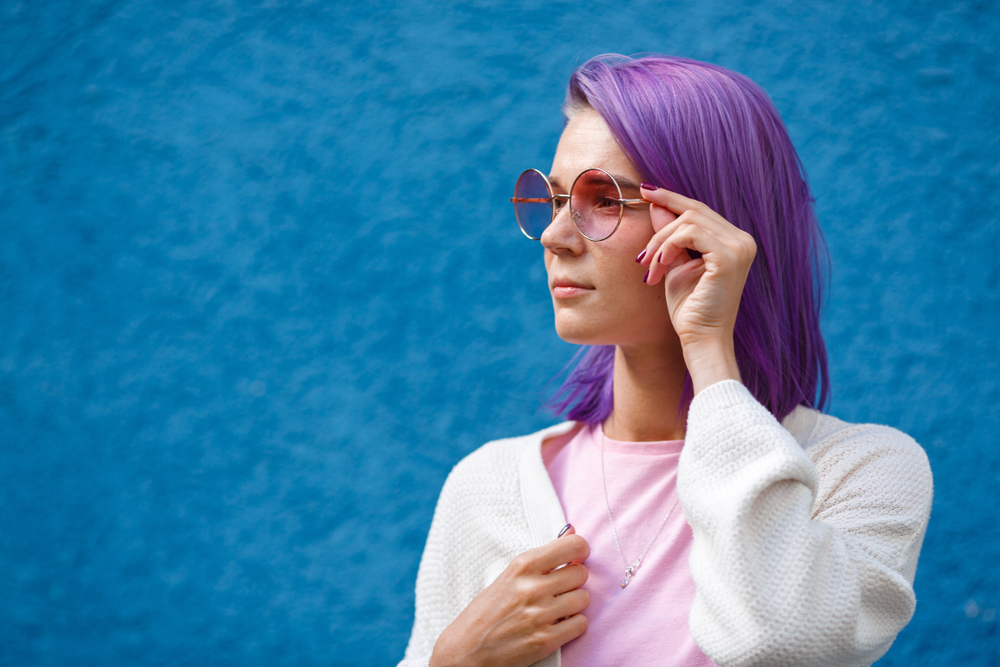 Vrouw in wit jasje en roze topje draagt grote ronde zonnebril met haar heldere lavendel lob kapsel voor een blauwe muur