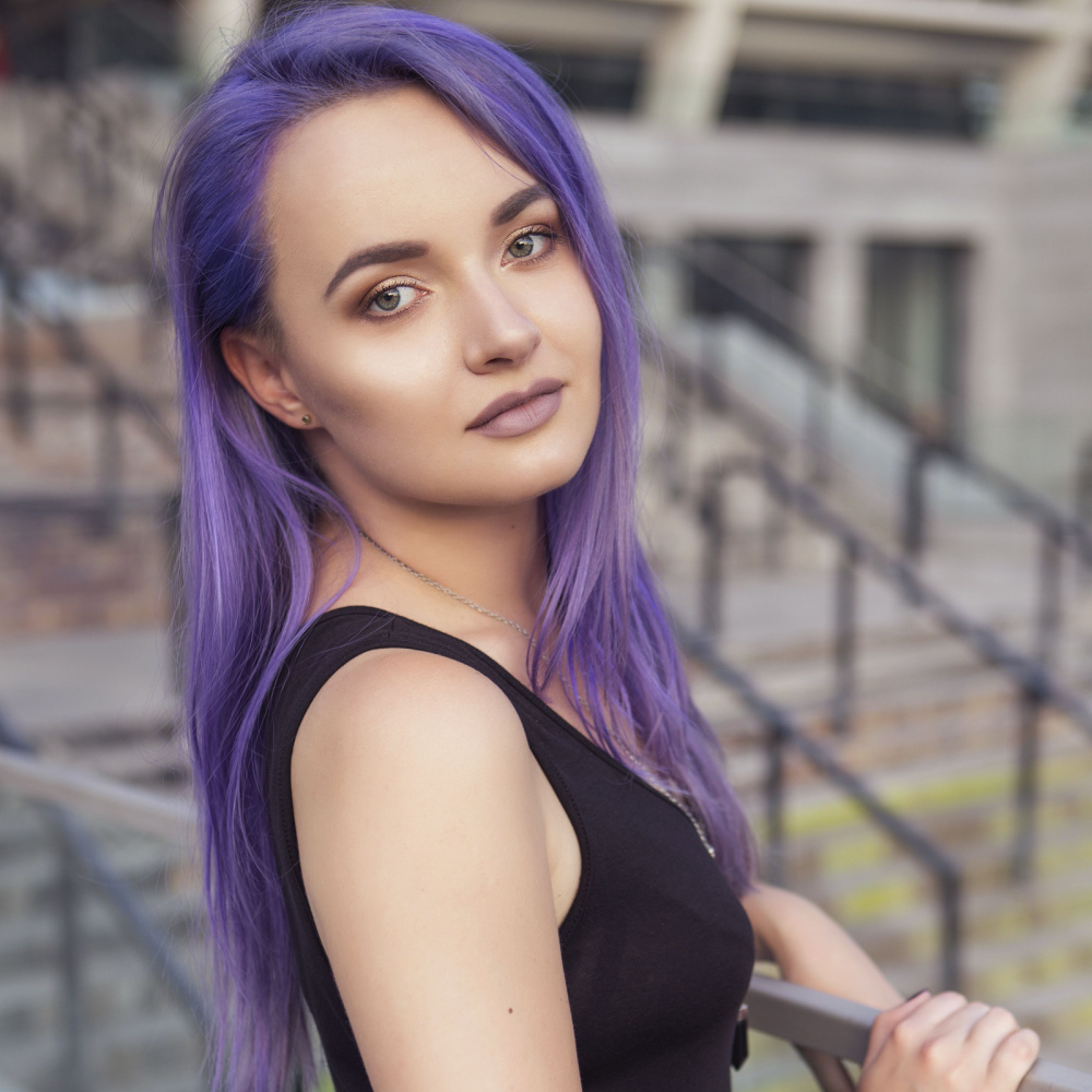 Vrouw met zwarte tanktop staat bij een reling in de stad met haar steile paarse lange haar gestyled met een zijscheiding