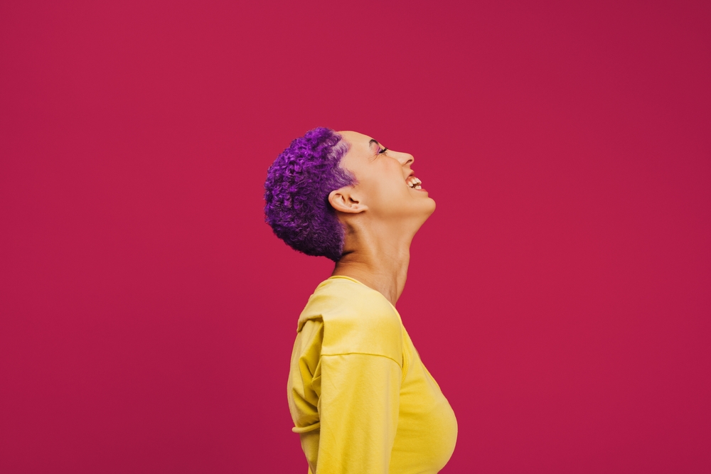 Vrouw met lavendel haar en medium huidskleur lacht met opgeheven hoofd en gesloten ogen terwijl ze een geel shirt draagt voor een rode achtergrond.