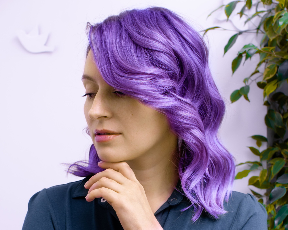 Close up van vrouw met hand onder haar kin die naar beneden kijkt met golvend schouderlang lavendelkleurig haar en groene bladeren tegen de muur achter haar.