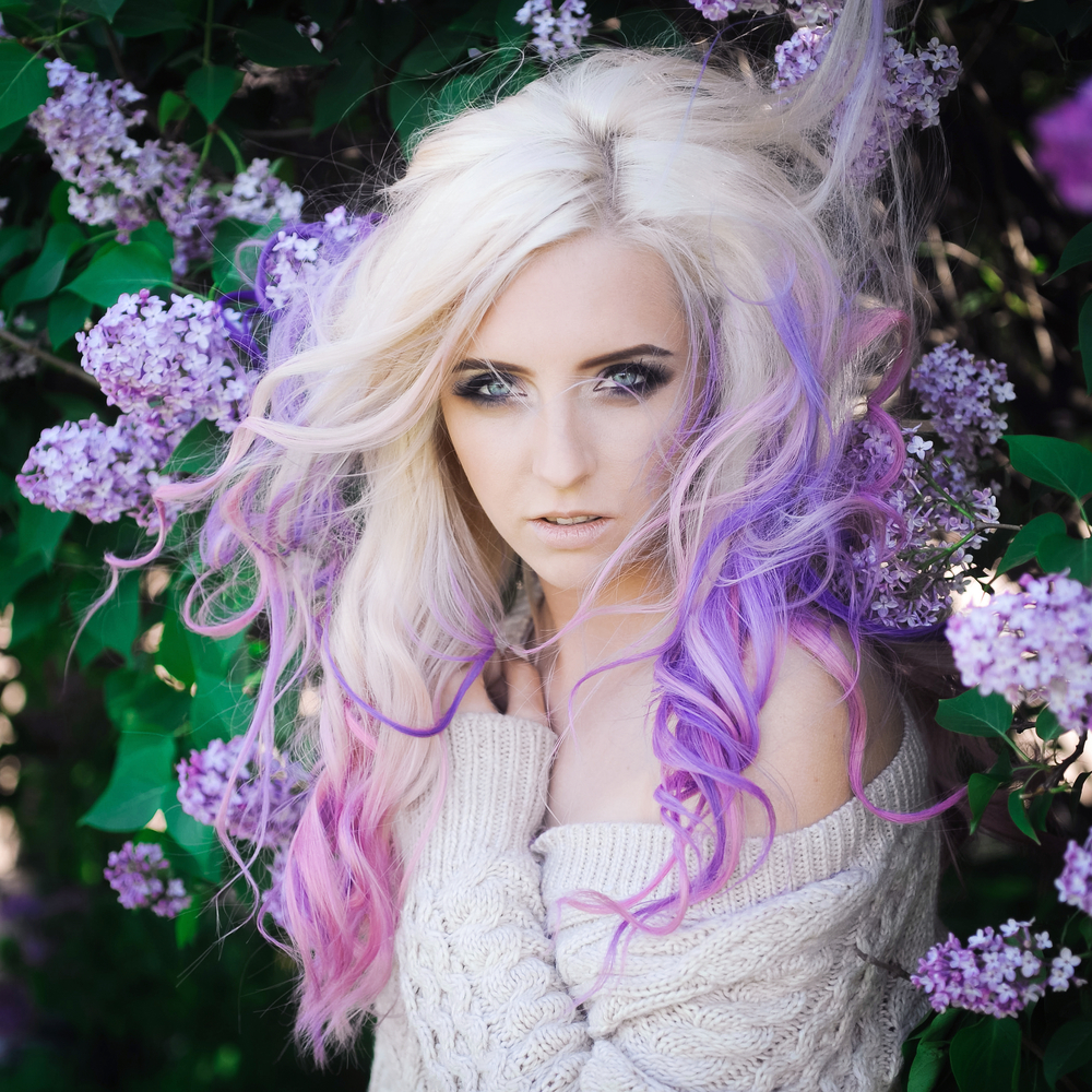 Blonde vrouw staat voor bloemstruiken met roze en lavendel haarkleuren en witte off-shoulder trui.