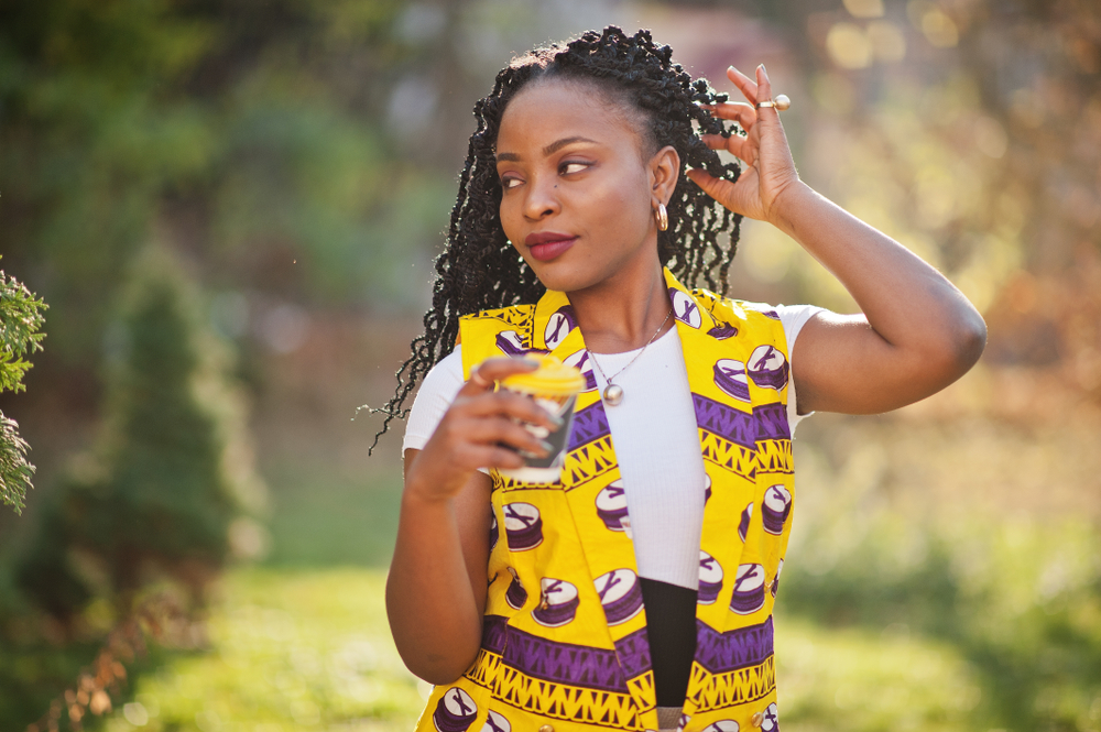 Gekleurde vrouw poseert met hand die haar spring twists achter haar oor plooit buiten met een glas water in een jurk met geel en paars patroon.