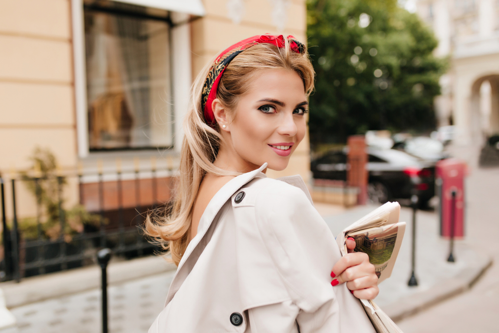 Blonde vrouw op een straat in de stad met jas over haar schouder kijkt glimlachend terug met rood hoofdbandkapsel voor vrouwen boven de 40