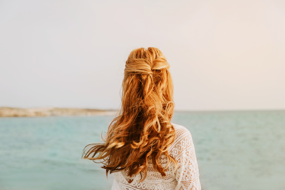 Achteraanzicht van een vrouw met koperrood haar die aan de kust staat met een half opgetrokken kapsel voor vrouwen van 40+ en een jurk met lange mouwen