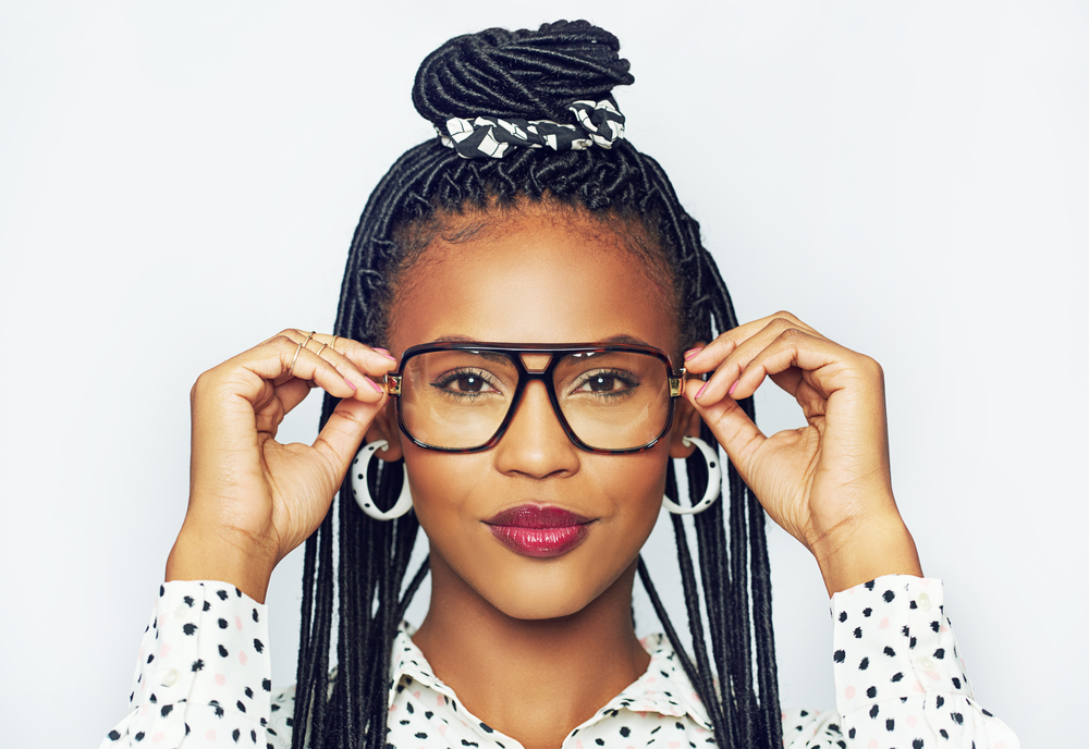 Vrouw zet een donker omrande bril op met haar faux locs en topknot kapsel met een wit stippenhemd en hoepeloorbellen 