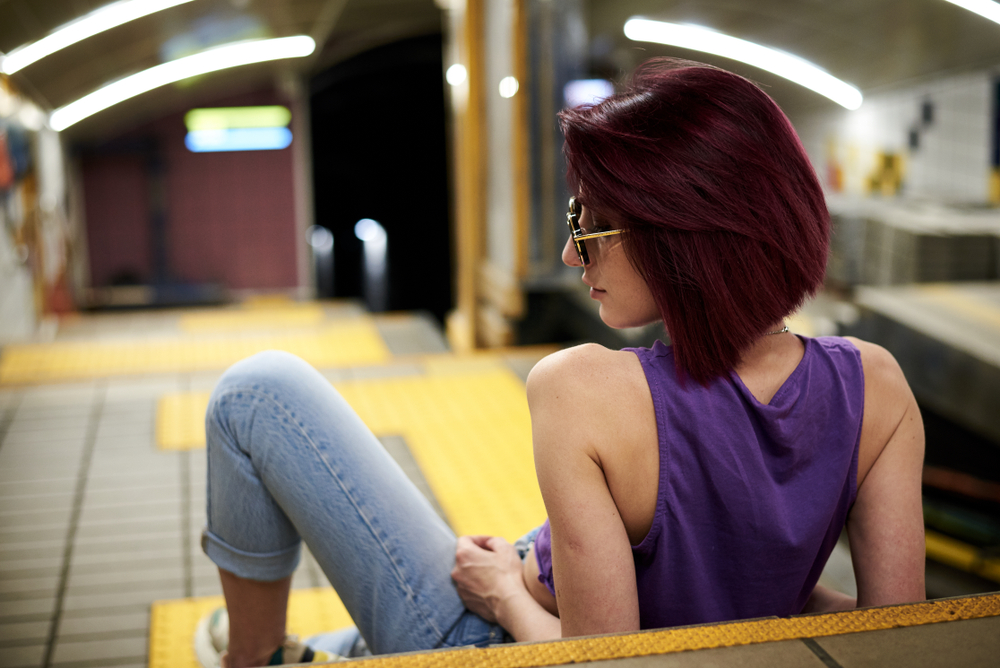 Vrouw leunt achterover op haar ellebogen in een metrostation om haar pruimkleurige haar en schuin bobkapsel te laten zien